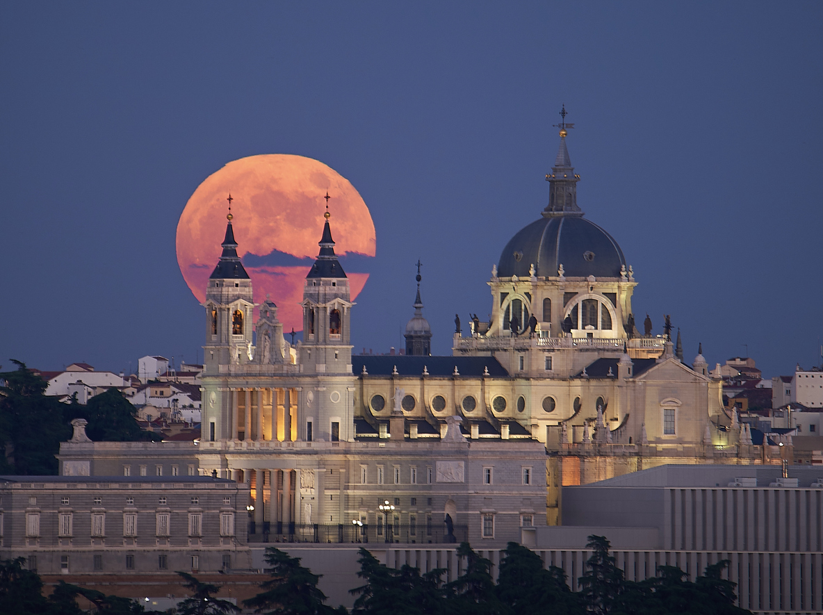 Full moon rising behind the illuminated Almudena Cathedral at twilight in Madrid, Spain. A stunning urban skyline with warm evening light and deep blue sky, a primary reason why Americans moving to Spain has become a thing.